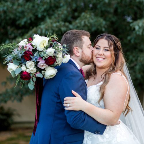 A young couple embrace at their wedding. The bride smiles for the camera, confident in her expertly styled hair and makeup.
